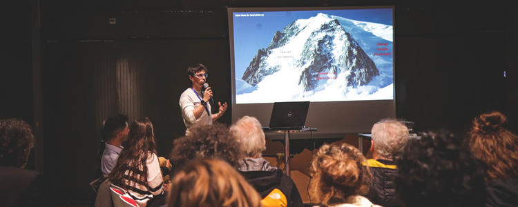 Le public écoute le conférencier, qui se tient devant la projection de son diaporama. Le diaporama montre une montagne enneigée